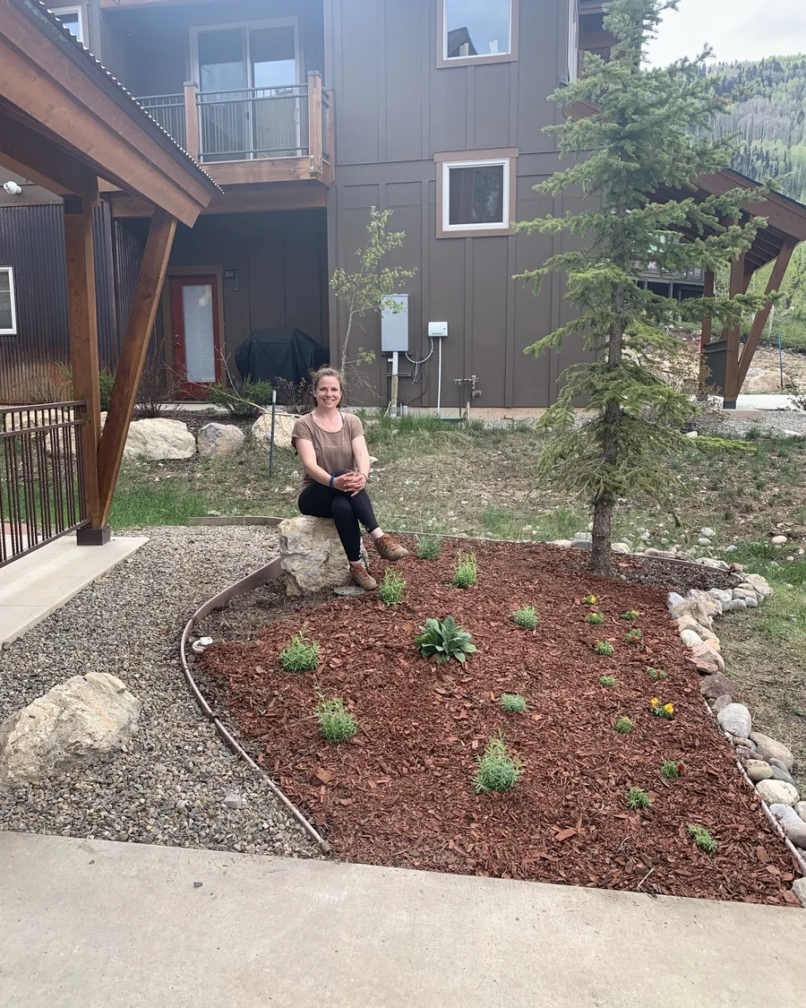 Katy seated beside a newly planted red-mulch garden bed around an evergreen in Durango, Colorado