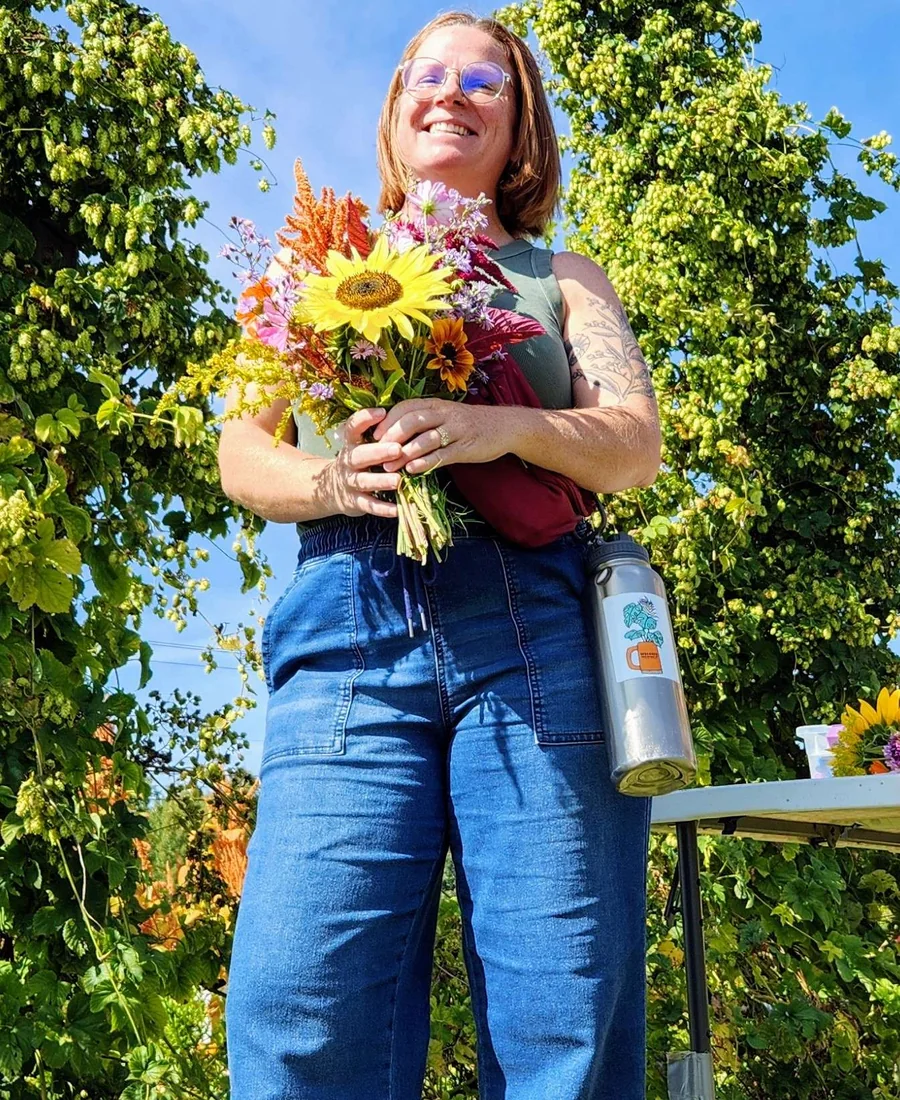 Katy Lydon McGreevy smiling and holding a bouquet of flowers beside growing hops